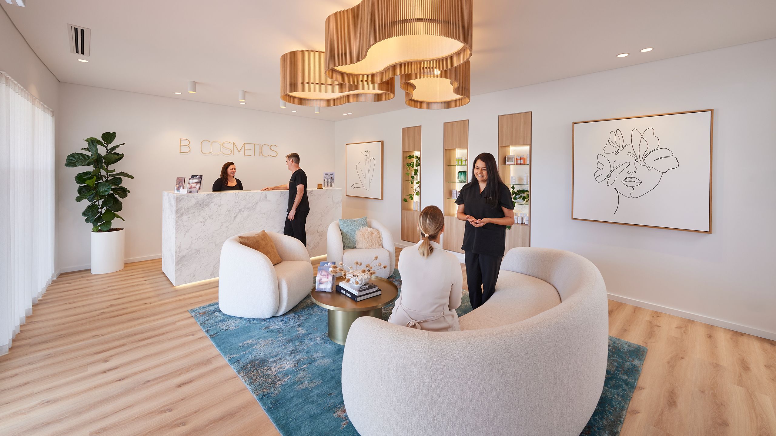 Inside B Cosmetics clinic, Dr Ben talks to the receptionist behind the counter, and Dr Sarah tals with a patient in the waiting room. The room is inviting and calm with plush furnishings and delicate artwork on the walls.