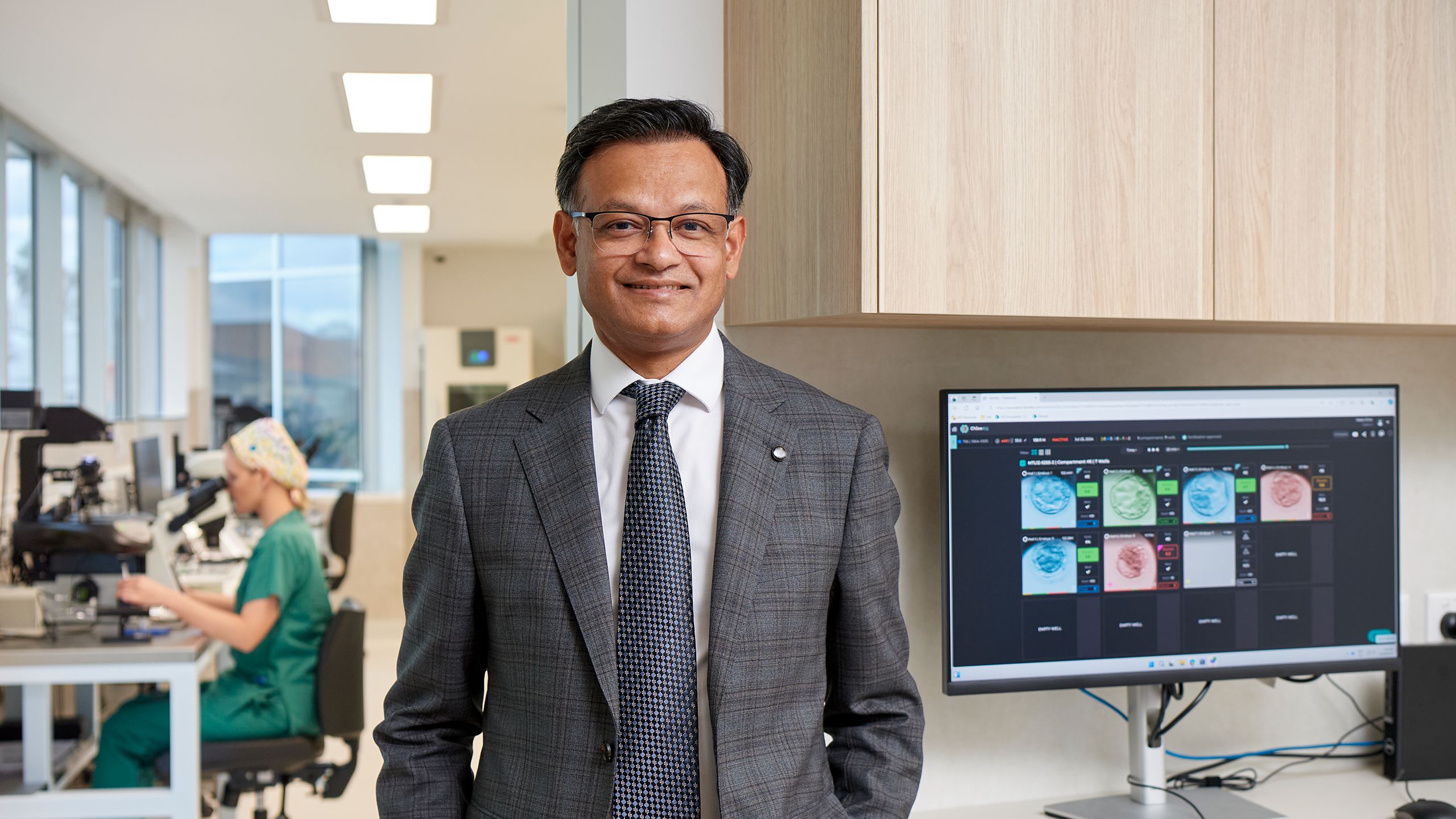 A profile image of Dr Santanu Baruah with a glimpse of the IVF lab and an embryologist in the background on the left and a computer showing scans of embryos on the right. 