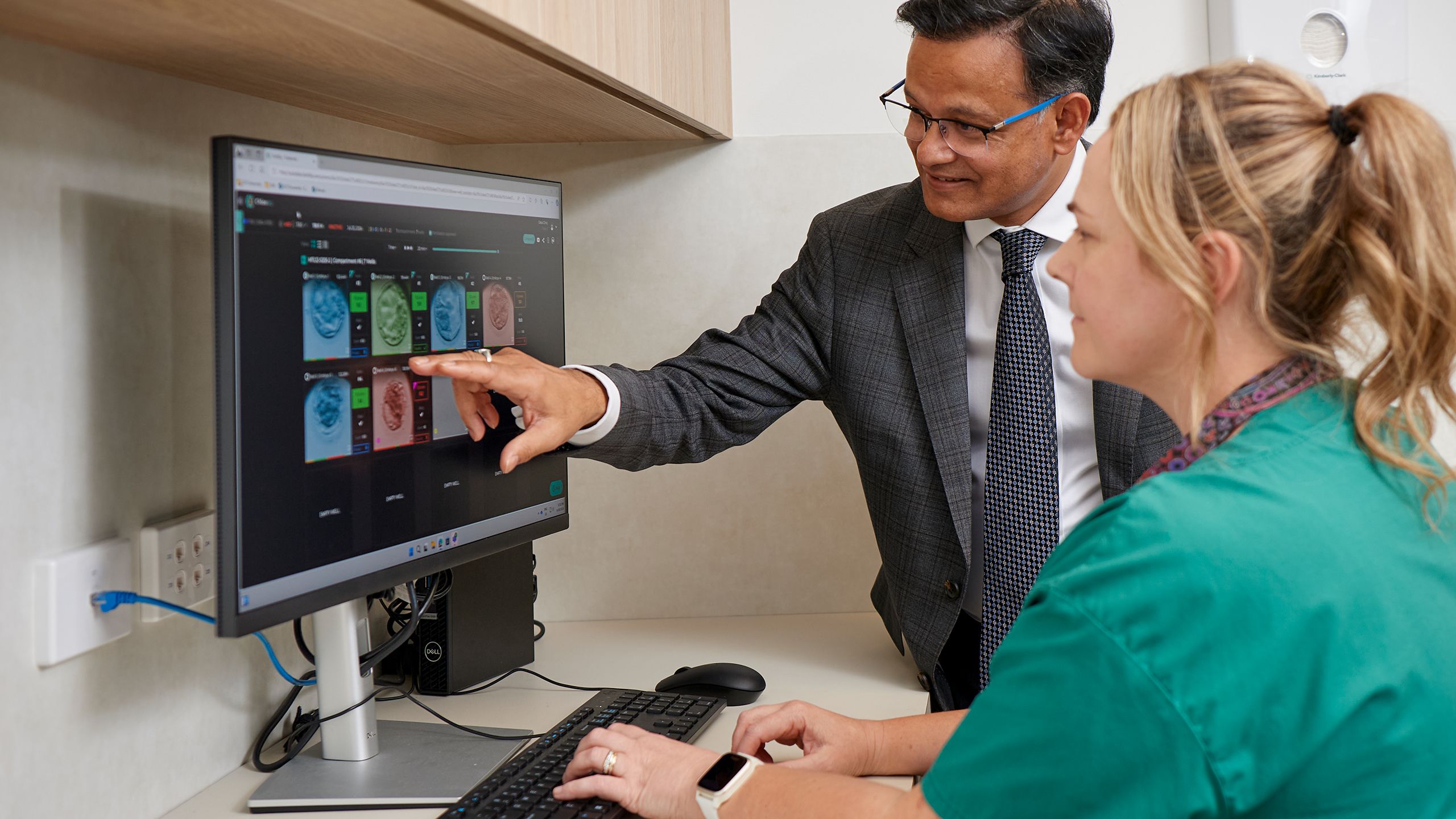 Dr Santanu Baruah points to a computer screen that shows embryos in different stages of growth. A woman in green scrubs sits in the foreground. 